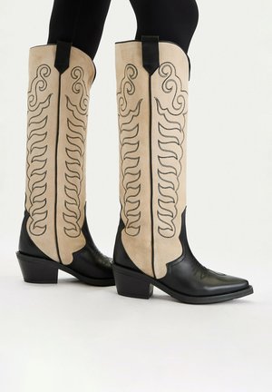 Wysokie kowbojki w beżowo-czarnym kolorze z przeszyciami w kształcie zawijasów, noszone z czarnymi leginsami, widoczne z boku na białym tle.