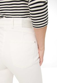 Hvide denimbukser med en tekstureret finish. Har en baglomme og et rektangulært mærke nær taljen med navnet "RAPHAELA."