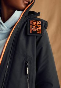 Jacket nera con zip e dettagli arancioni, tessuto texturizzato e una vistosa toppa del marchio. Presenta un colletto alto e tasche laterali con zip.