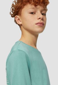 Młoda osoba z rudymi włosami i piegami, ubrana w jasnoniebiesko-zieloną koszulę, patrzy pewnie przez ramię na tle jednolitego tła.