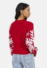 Pull rouge tricoté avec des motifs de flocons de neige blancs sur les manches et un ourlet côtelé. Il présente un col rond et une coupe légèrement raccourcie.