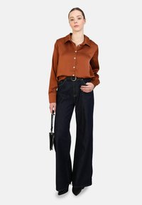 Braune Satin-Hemdbluse mit langen Ärmeln, kombiniert mit hochtaillierten, dunkelblauen Jeans im weiten Schnitt. Ein schwarzer Gürtel und eine kleine Handtasche setzen Akzente.