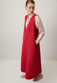 Červené vlněné šaty bez rukávů s hlubokým výstřihem do V, texturovaným povrchom, bočnými vreckami a voľným strihom, skombinované so svetloružovou košou a topánkami.