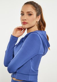 Felpa blu con zip, dal design aderente, maniche lunghe e cappuccio piccolo. Realizzata in tessuto liscio con una leggera lucentezza e dettagli di cucitura.
