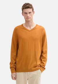 Suéter de cuello en V en un color naranja quemado, hecho de una tela suave. Presenta mangas largas y un dobladillo y puños acanalados. Diseño sencillo y clásico.