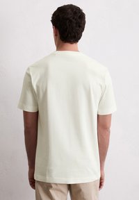 Bărbat îmbrăcat cu un tricou simplu alb-crem cu mânecă scurtă și pantaloni bej, stând cu fața spre un perete texturat alb.