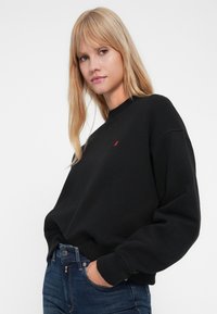 Svart hettegenser laget av mykt materiale, med rund hals, senket skuldre og en liten rød logo på brystet. Brukt sammen med blå jeans.
