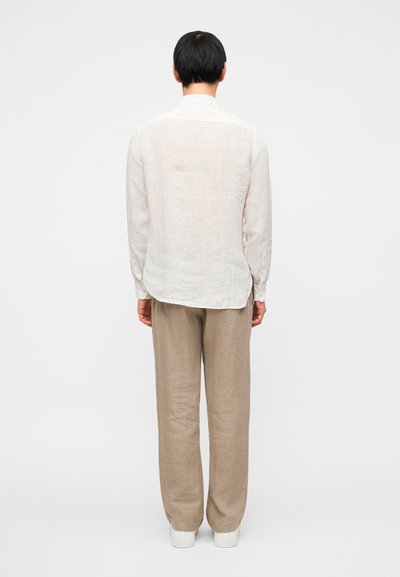 Λιτή πουκαμίσα από λινό σε ανοιχτό μπεζ χρώμα με μακριά μανίκια και γιακά με κουμπιά, συνδυασμένη με άνετο, ανοιχτό καφέ λινό παντελόνι και λευκά αθλητικά παπούτσια.