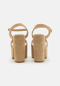 Talons compensés en suédine beige avec un talon bloc épais, une lanière à la cheville et un dos ouvert. Texture lisse avec un design à bout rond.