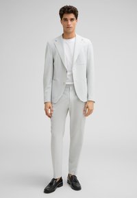 Un costume gris clair texturé avec un blazer à un bouton, un pantalon assorti et un t-shirt blanc. Des chaussures noires complètent l'ensemble.