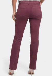 Jeans elásticos burdeos con corte ajustado, cintura media, dos bolsillos traseros y costuras expuestas; la modelo lleva puestos tacones beige.