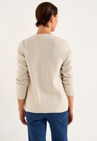 Ljust beige stickad tröja med ett texturerat mönster, rundad hals och trekvartsärmar, kombinerad med blå denimjeans.