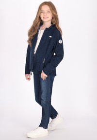 Giacca zip-up blu navy in morbido tessuto con colletto alto, dotata di una toppa con logo sulla manica, abbinata a jeans di denim scuro e sneaker bianche.