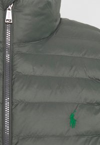 Puffjacka i mörkgrönt med kviltad textur, med dragkedja fram och en broderad logotyp i ljusgrönt på bröstet.