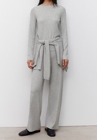 Conjunto de punto gris compuesto por un top de manga larga con cinturón en la cintura y pantalones anchos a juego, ambos con una sutil textura acanalada. El look se completa con zapatos planos negros.