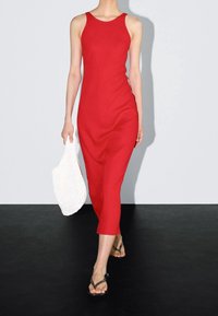 Femme marchant dans une robe midi rouge sans manches, tenant un sac blanc texturé dans une main, portant des sandales noires à lanières sur un sol noir.