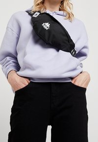 Czarna saszetka na biodra z logo, noszona ukośnie na lawendowej bluzie z kapturem. Miękki materiał, smukły kształt, minimalistyczny design. Czarne spodnie z kieszeniami.