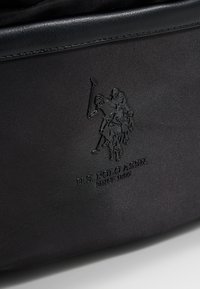 Černá tkaninová taška s vystouplým logem jezdce na koni s polovým koulí, detailním prošíváním a textem "U.S. POLO ASSN. SINCE 1890."