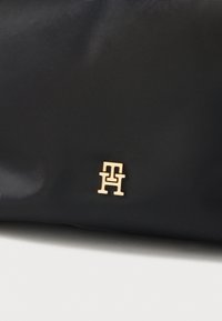 Geantă neagră din nylon, cu un emblemă de logo auriu formată din litere interconectate. Textură netedă, design minimalist, fără alte modele.