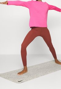 Rosa långärmad topp med avslappnad passform och sidospalter, matchad med bruna högmidjade leggings. Står på en ljusgrå mönstrad yogamatta.