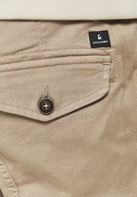 Poche beige de pantalon avec un rabat boutonné et une étiquette noire de la marque Jack & Jones au-dessus de la poche.