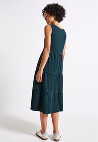 Teal sleeveless dress with a tiered design. Made from soft fabric, knee-length, featuring a small button detail at the back. Paired with casual shoes.