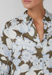 Blusa floreale con grandi motivi di fiori blu e bianchi su uno sfondo marrone, design con bottoni e colletto classico.
