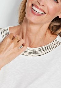 Weißes Oberteil mit texturiertem Finish und einem Halsausschnitt, der mit goldenen Nieten verziert ist. Eine Hand ruht nah am Kragen und präsentiert einen goldenen Ring.