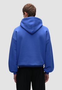 Niebieska bluza z kapturem o luźnym kroju, przestronnym kapturze i miękkiej fakturze. Posiada opadające ramiona oraz skróconą długość, zaprezentowana od tyłu.