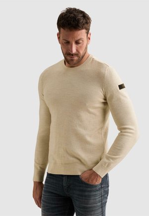 Man wearing a beige long-sleeve sweater with a logo patch on the left arm and dark jeans, looking down with one hand in pocket.