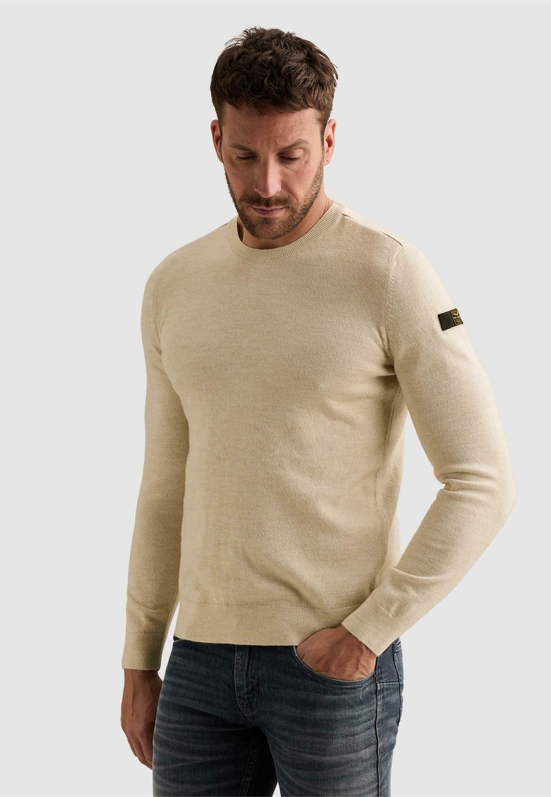 Homme portant un pull beige à manches longues avec un patch logo sur le bras gauche et un jean foncé, regardant vers le bas avec une main dans la poche.