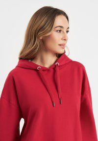 Rode pullover hoodie met een voorkant kangaroo zak, koordjes en zijsplitten. Gemaakt van een zachte katoenmix, heeft een relaxte pasvorm en een capuchon.