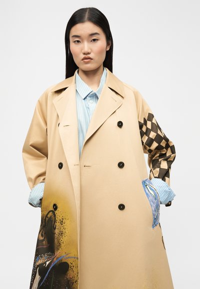 Γυναίκα με μακριά σκούρα μαλλιά που φοράει ένα μπεζ παλτό καμπαρντίνα με μαύρο καρό σχέδιο σε ένα μανίκι και πολύχρωμα αφηρημένα σχέδια.