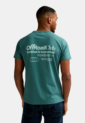 Mann trägt ein türkisfarbenes OffRoadClub-T-Shirt mit weißem Aufdruck und Logo, blickt mit dem Rücken zur Kamera vor einem schlichten hellen Hintergrund.