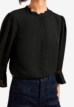 Femme portant un chemisier noir à manches longues avec des épaules bouffantes et un col cranté, rentré dans un jean en denim foncé taille haute.