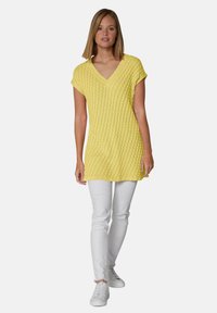 Gelbe strukturierte Tunika mit V-Ausschnitt und kurzen Ärmeln, kombiniert mit weißen Slim-Fit-Hosen und weißen Sneakers.