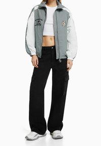 Šedá a bílá zipová bunda s logem, kombinovaná s volnými černými cargo kalhotami. Model má na sobě bílé tenisky. Neformální, sportovní outfit.