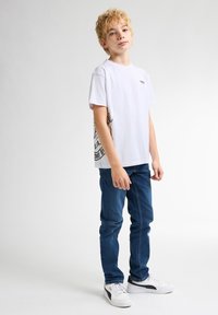 Bílý tričko s krátkým rukávem a černou grafikou na boku, spárované s modrými úzkými džíny a bílými teniskami s černými akcenty.