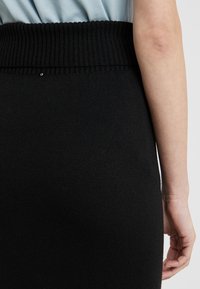 Jupe en tricot noire avec une taille côtelée. La texture est lisse, présentant un léger éclat et un petit détail décoratif près de la taille.