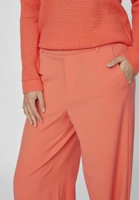 Žena v korálově barevném texturovaném svetru a odpovídajících širokých kalhotách, s jednou rukou odloženou v kapse a druhou u boku.