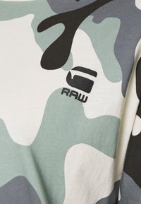 T-paita lyhyillä hihoilla, jossa on kamouflagekuvio erivärisin vihrein, harmaisin ja valkoisin sävyin; edessä musta logo "RAW".