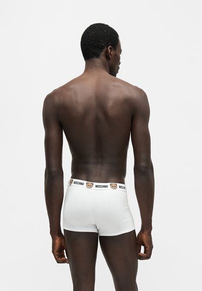 Modèle masculin adulte vu de dos portant un boxer blanc Moschino avec logo ours en peluche sur la ceinture, sur fond blanc uni.