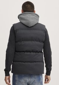 Chaleco acolchado gris oscuro con cuello alto, llevado sobre una sudadera con capucha gris. Presenta puños acanalados y paneles laterales de punto texturizado.