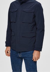 Marinblå jacka i lätt tyg, med hög krage, fyra framfickor och justerbara ärmslut. Bärs över denimjeans.