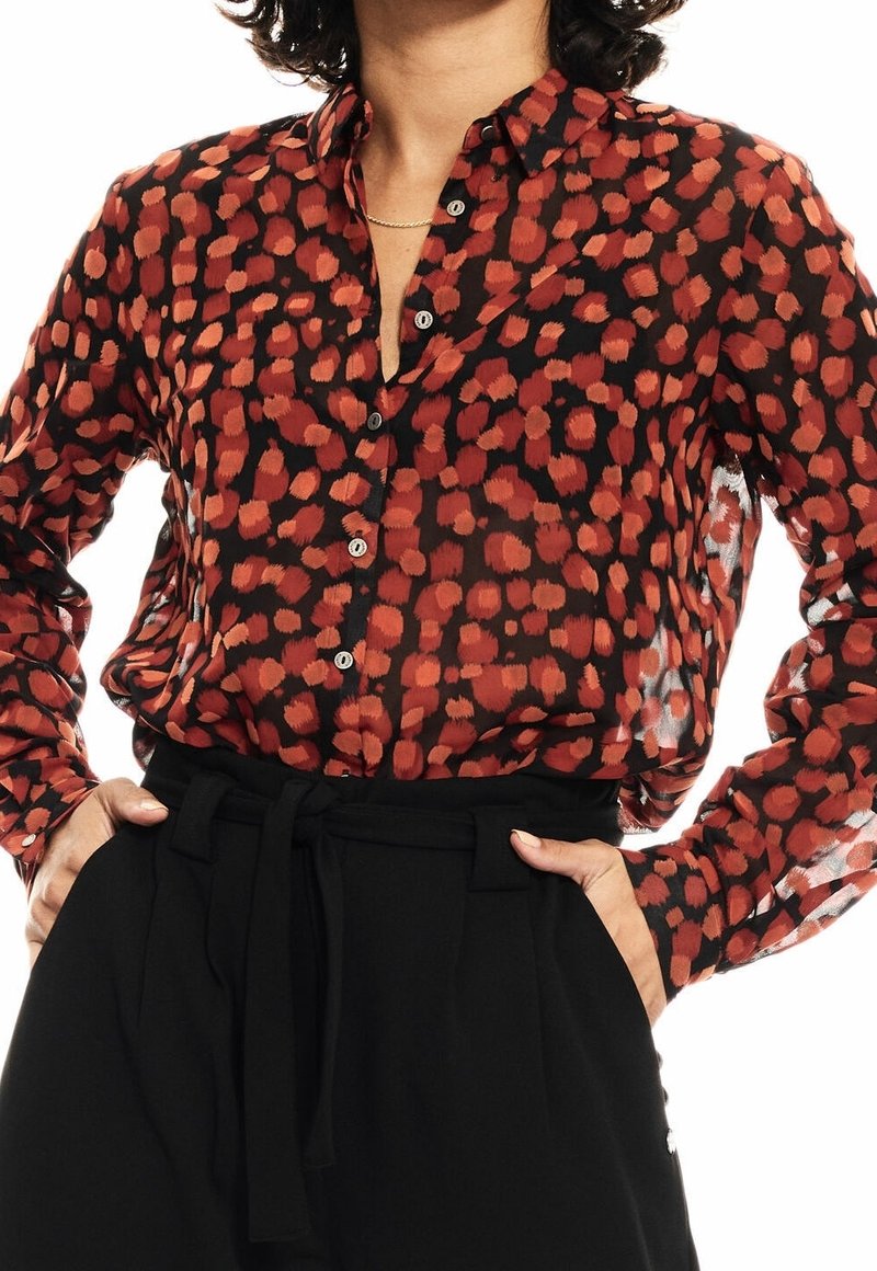 Czerwona i czarna wzorzysta bluzka z zapinanym na guziki przodem, półprzezroczystym materiałem i długimi rękawami. W połączeniu z czarnymi, wysokim stanem spodniami.