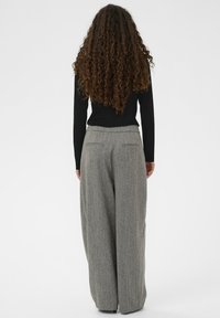 Sieviete ar gariem cirtainiem matiem, tērpta melnā garajās piedurknēs un pelēkās plašās biksēs, stāv, atspiedusies pret vienkāršu fona.