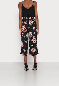 Svart blommig jumpsuit med breda cropped ben, prydd med livliga mönster av rosa, vita och gröna blommor; tillsammans med svarta högklackade sandaler.