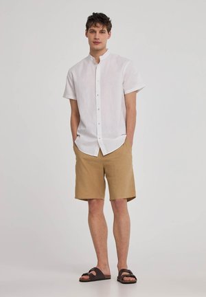 Camicia bianca di lino a maniche corte con colletto alla coreana, abbinata a pantaloni corti di lino color sabbia e sandali marroni scuri, in piedi davanti a uno sfondo neutro.