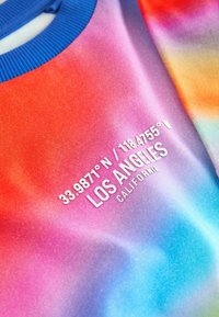 Kurzarmpullover mit einem Farbverlauf in roten, rosa und blauen Farbtönen. Mit weißem Text auf der Vorderseite, der die Koordinaten und "LOS ANGELES" zeigt.