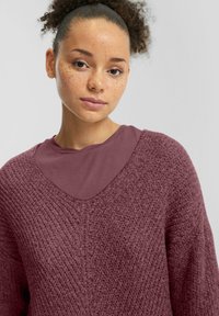 Pull en maille texturée bordeaux avec un décolleté en V profond et un top bordeaux superposé en dessous. Tissu doux avec des détails de couture visibles.